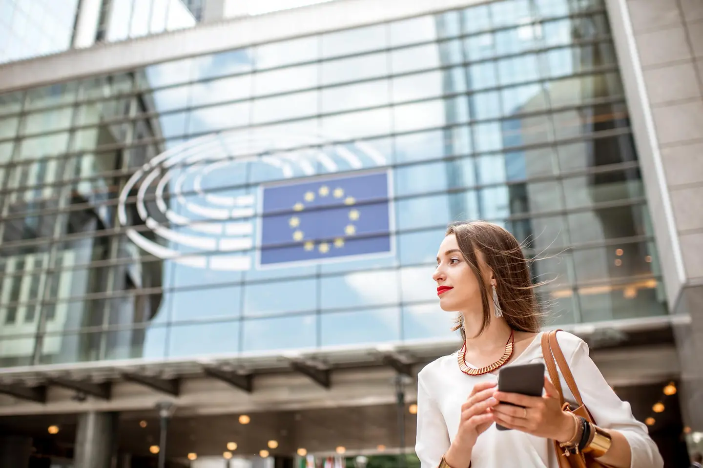 Junge Geschäftsfrau mit Smartphone steht nahe dem Parlamentsgebäude der Europäischen Union in Brüssel